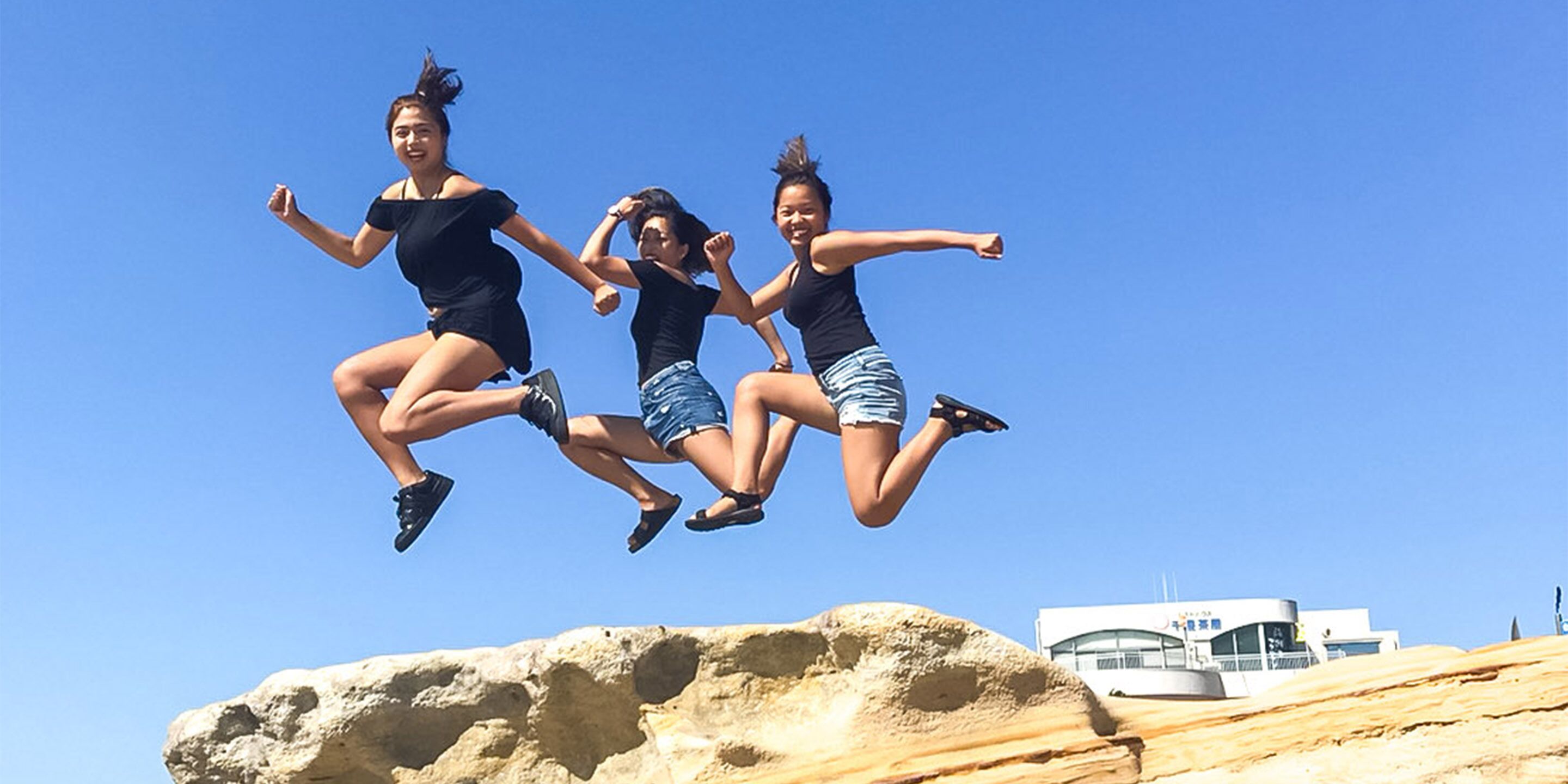 Three people jumping off a rock with a building in the background.