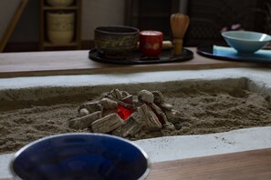 A close-up of a charcoal fire with wood logs and ash, surrounded by a sandy area. - Guest Cafe Kuchikumano (Kamitonda)