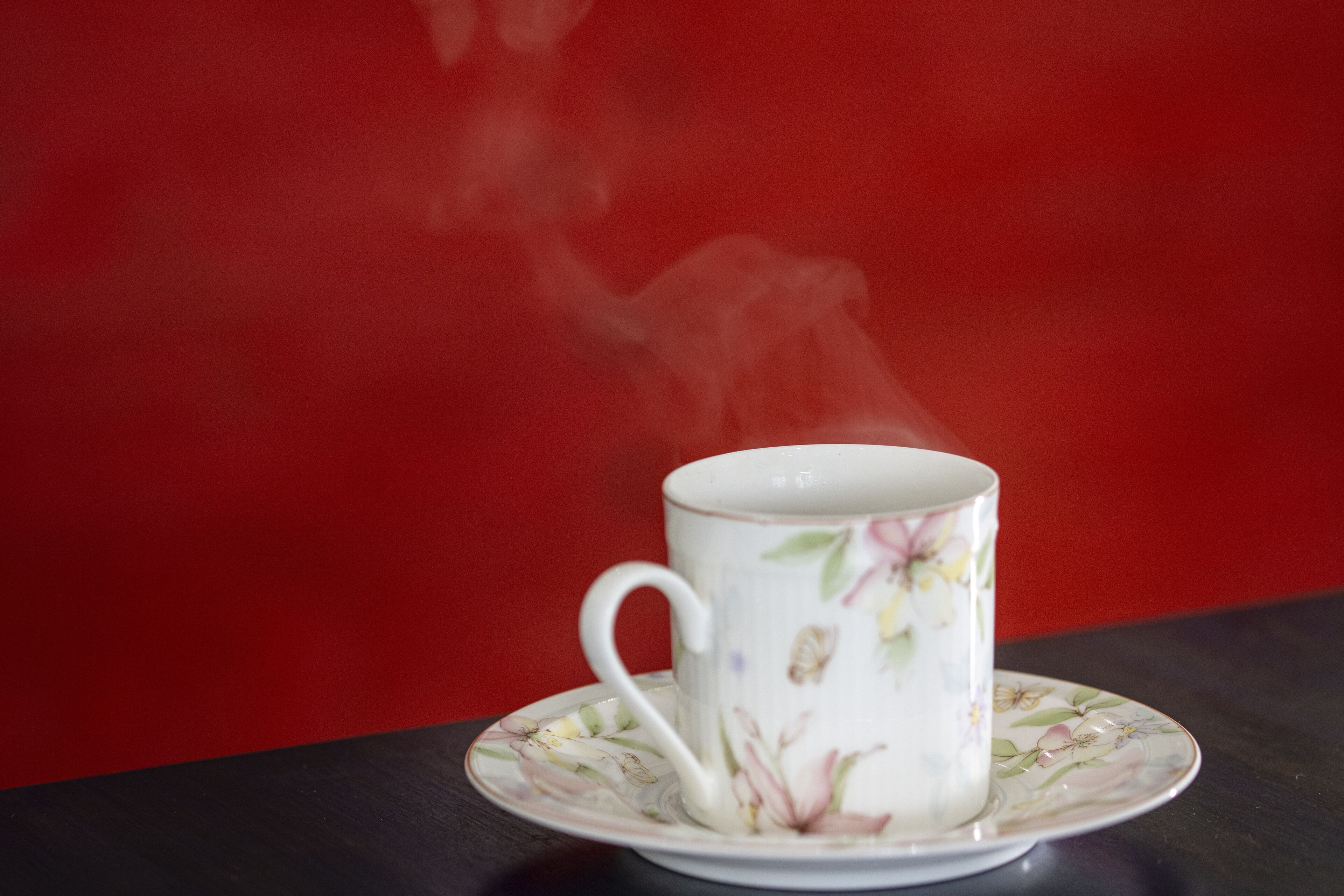 A white ceramic cup with floral designs on a matching saucer, placed on a dark surface against a red background.