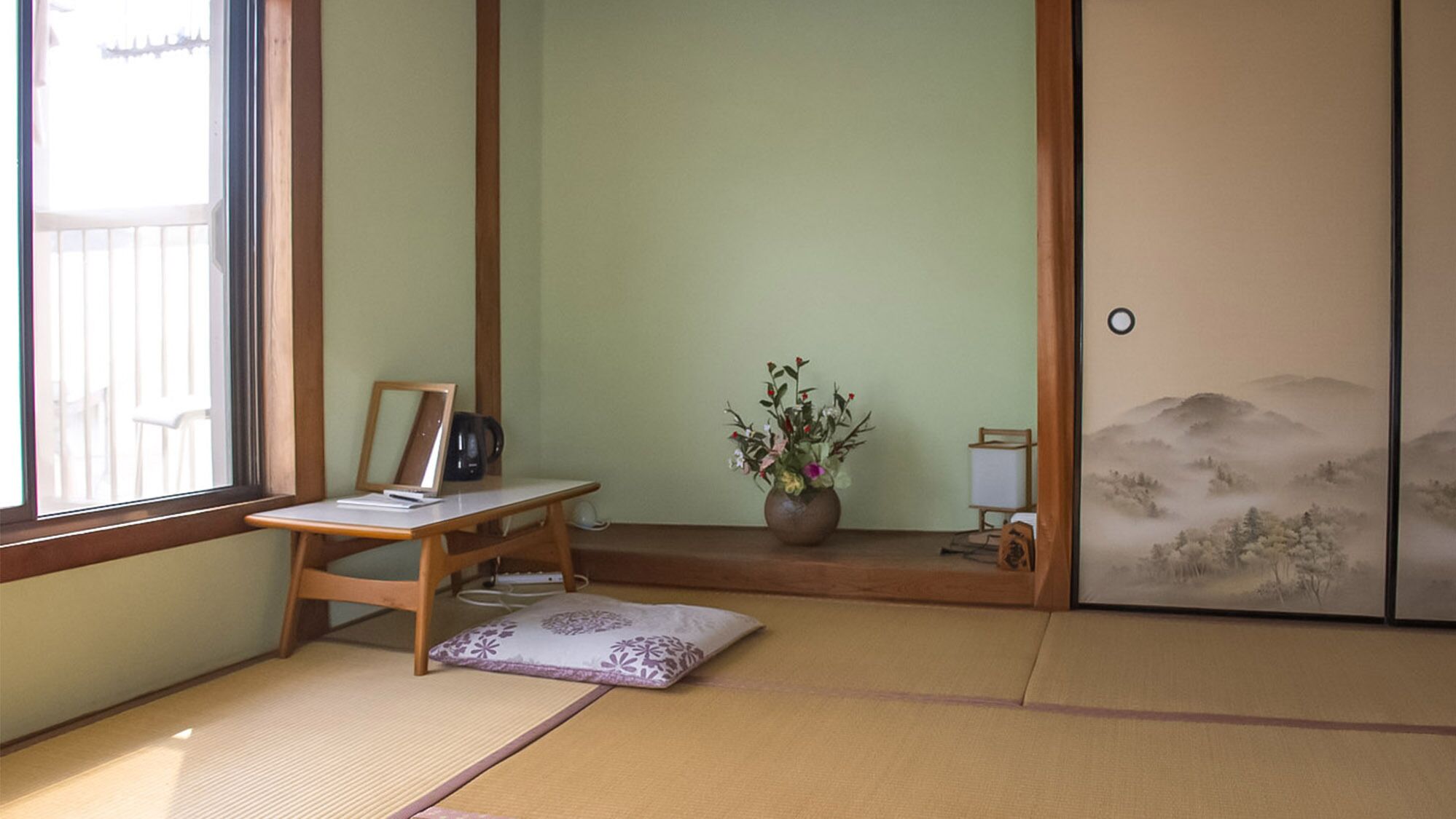 A traditional Japanese room with tatami flooring, a low table with a picture frame, a vase with flowers, and a sliding door with a landscape painting.
