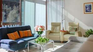 Ein Wohnzimmer mit einem blauen Sofa, beigen Sesseln, einem Glastisch und einem großen Fenster mit Vorhängen.