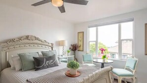 Une chambre avec un grand lit, un ventilateur de plafond, une fenêtre avec vue, une petite table avec une lampe et une chaise.