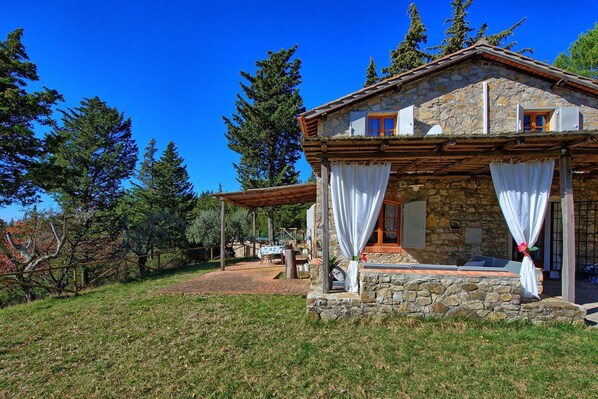 A stone house with a covered porch, a dining area with outdoor seating, and a view of surrounding trees.