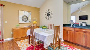 A dining area with a table set for four, wooden chairs, a decorative wall piece, and a kitchen with wooden cabinets and a black countertop.
