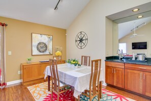 A dining area with a table set for four, wooden chairs, a decorative wall piece, and a kitchen with wooden cabinets and a black countertop. - Close to Rehoboth & Dewey w/ Access to 3 Pools! (Rehoboth Beach)