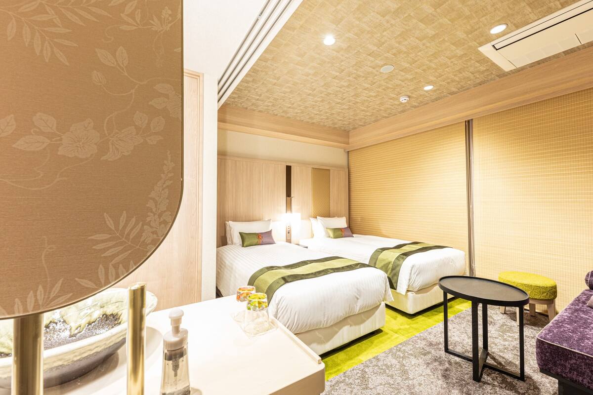 Ein Hotelzimmer mit zwei Betten, einem kleinen Tisch, einer Couch und einem Badezimmer mit Spiegel und Pflegeprodukten.