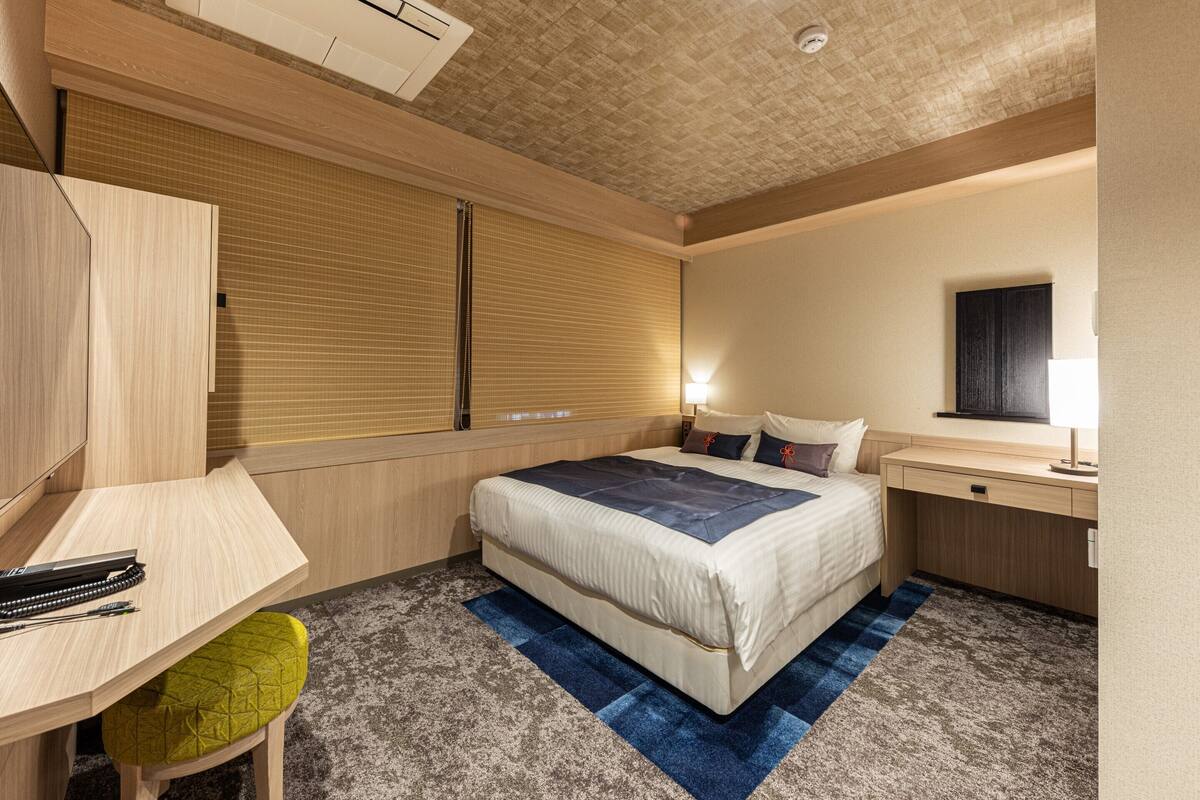 Ein Hotelzimmer mit Bett, Schreibtisch und Stuhl, einem Fenster mit Jalousien, einer Nachttischlampe und einer Klimaanlage.