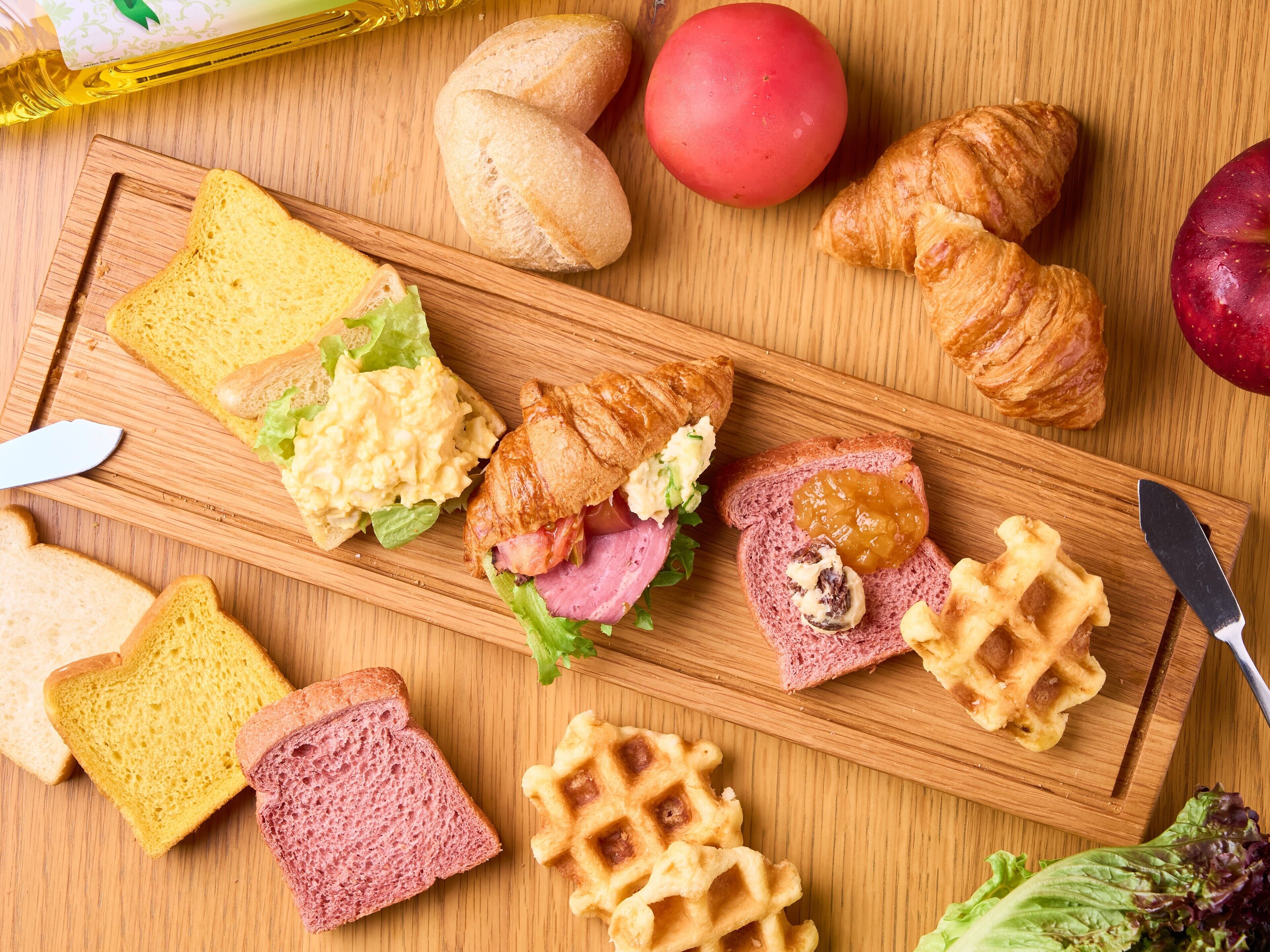 A wooden cutting board with various food items including bread, croissants, a tomato, and a waffle.