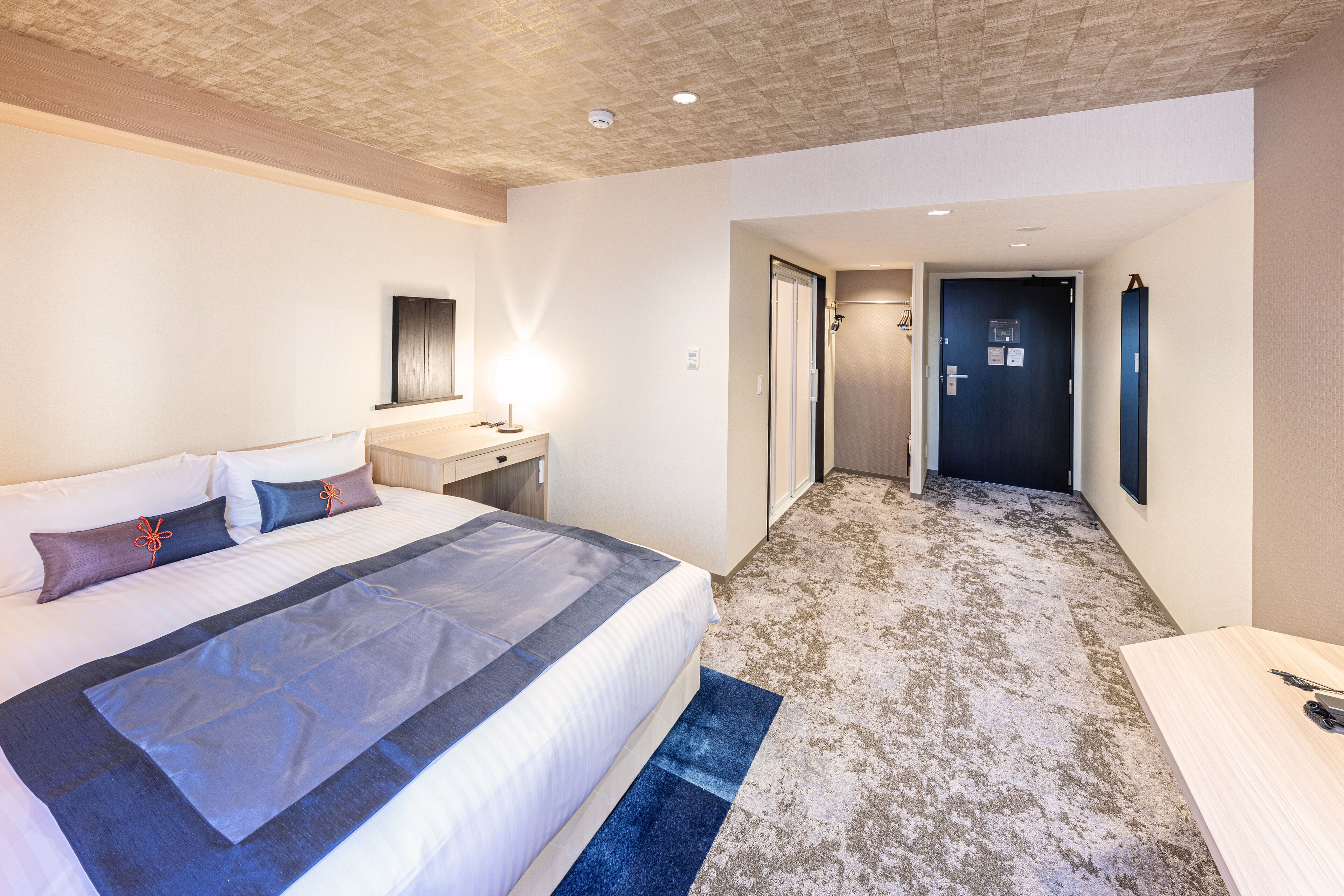 A hotel room with a bed, a desk, a mirror, and a door leading to a hallway.