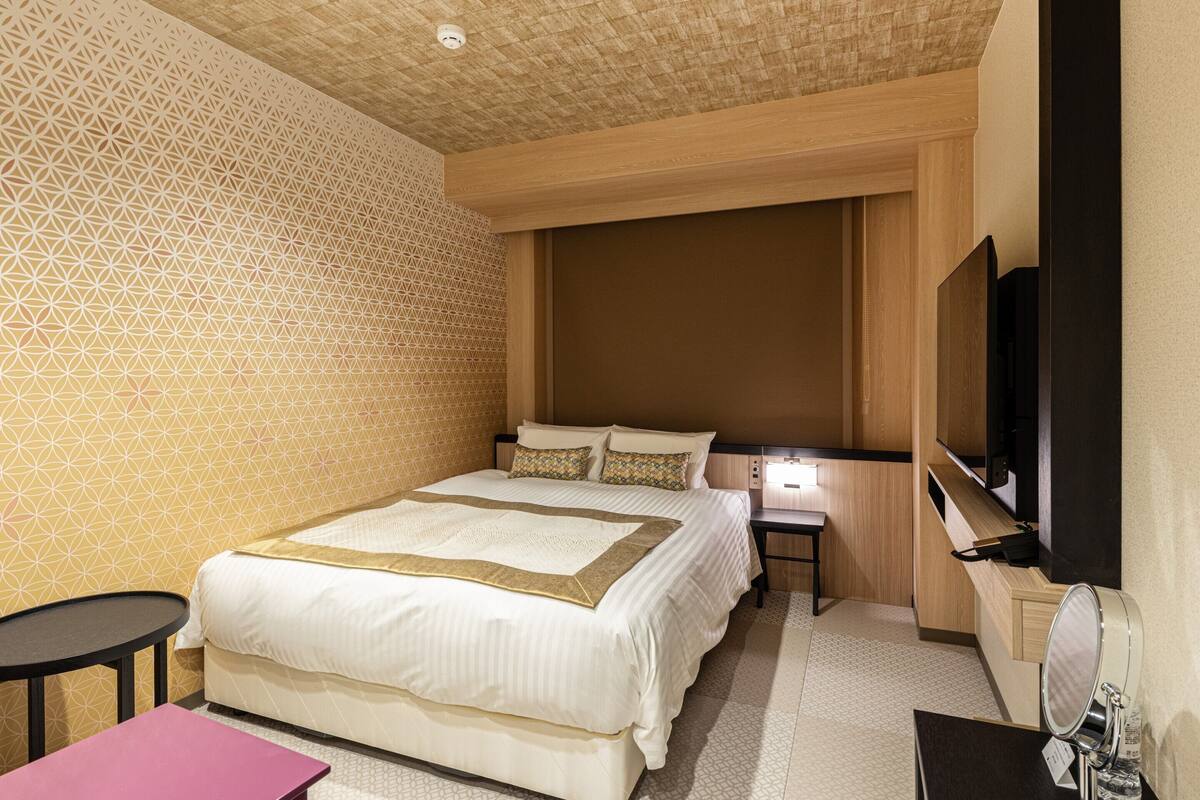 Ein Hotelzimmer mit einem großen Bett, einem kleinen Tisch, einem Nachttisch mit Lampe und einem an der Wand befestigten Fernseher.