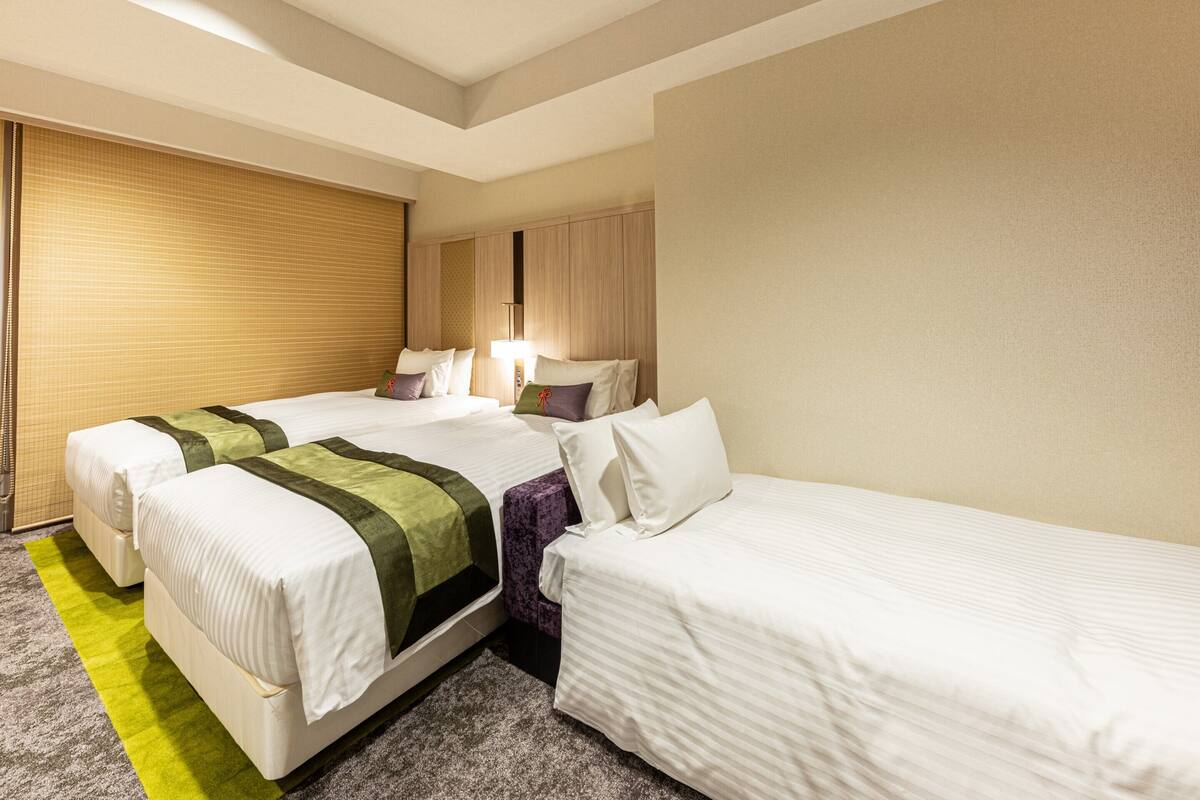 Ein Hotelzimmer mit zwei Betten, einem großen Fenster mit Jalousien, Teppichboden und einer Deckenleuchte.