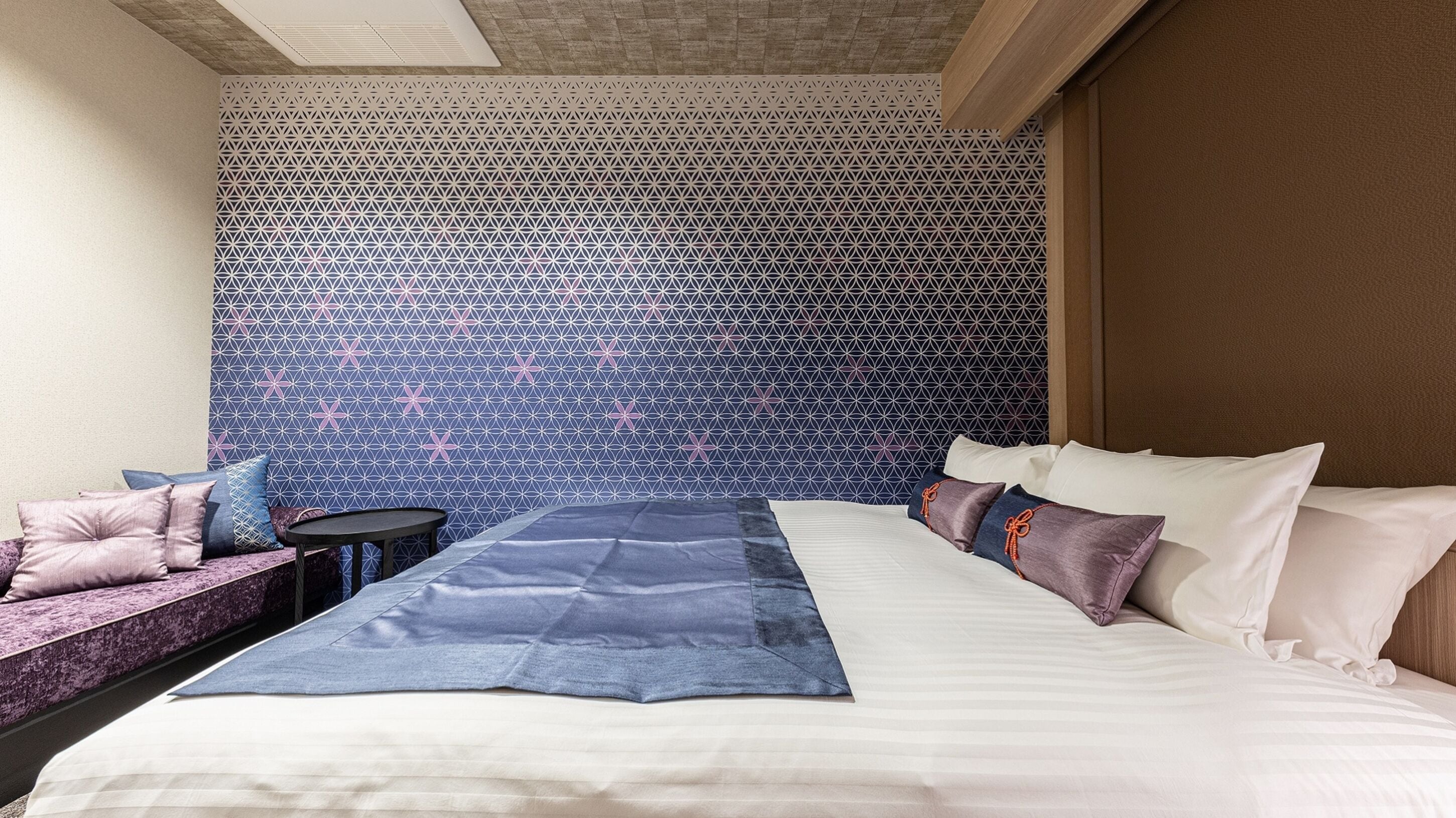 A hotel room with a bed, a small table, a bench, and a patterned wall.
