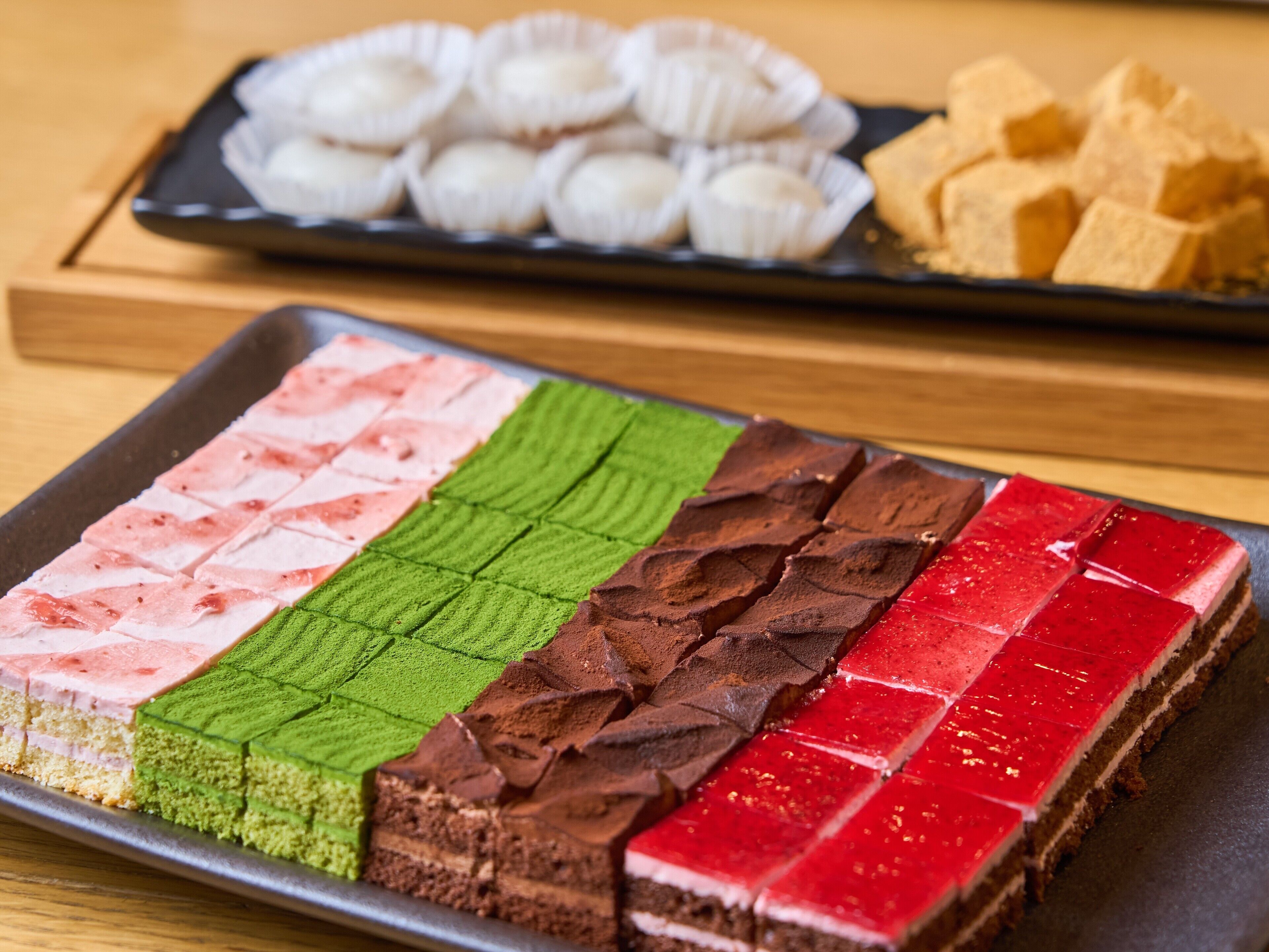 Una bandeja de postres con distintas capas coloridas y una fuente de cupcakes al fondo.