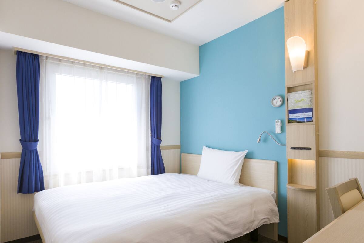 A hotel room with a large bed, a desk, a chair, a nightstand, a wall clock, and a wall-mounted lamp.