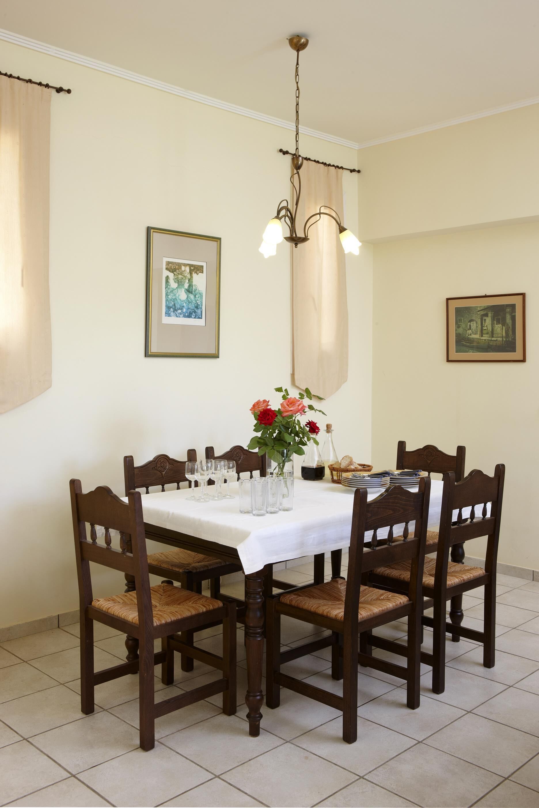 A dining room with a white table, six wooden chairs, a chandelier, and two framed pictures on the walls.