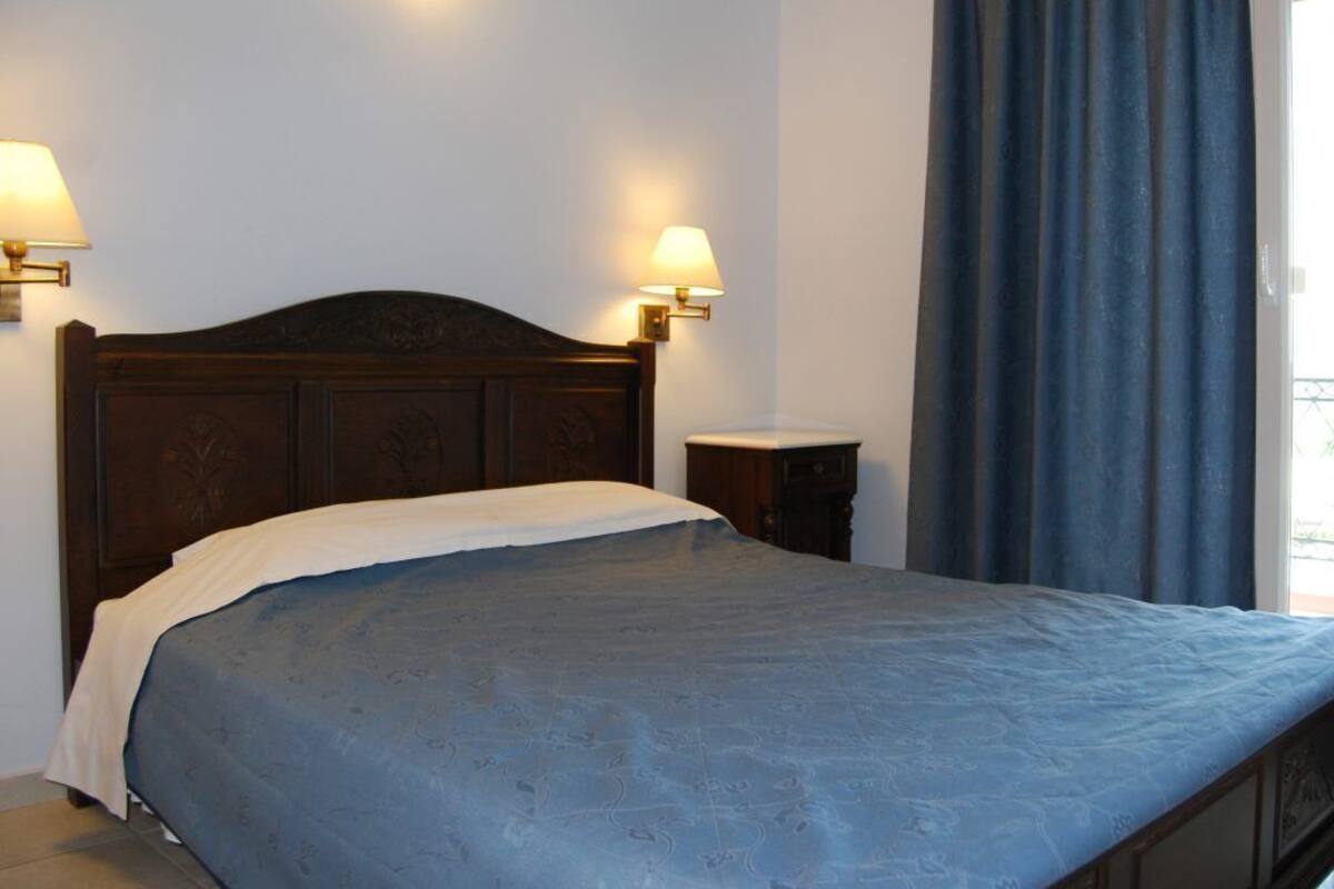 Une chambre à coucher avec une tête de lit en bois, un lit recouvert d’une couverture bleue, deux lampes fixées au mur, une table de chevet et une fenêtre ornée de rideaux bleus.