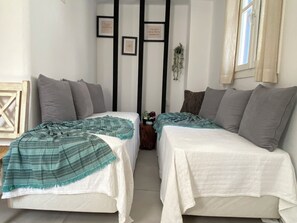 Una habitación con dos camas, cada una con una colcha blanca y una manta verde azulada. Hay cuadros enmarcados en la pared y una planta pequeña en una mesita de noche.