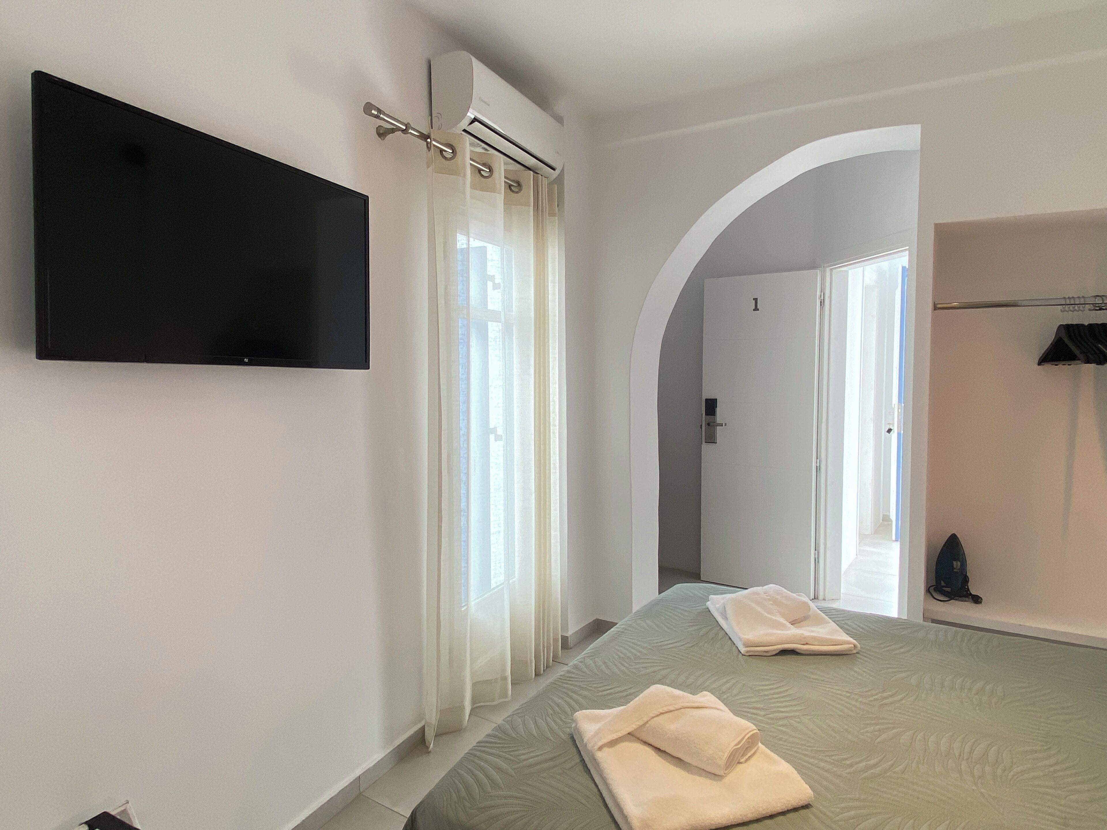 Ein modernes Schlafzimmer mit einem an der Wand befestigten Flachbildfernseher, einem Bett mit grüner Bettdecke, zwei gefalteten Handtüchern und einem begehbaren Kleiderschrank mit Kleiderstange.