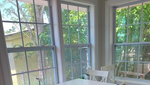 A dining area with a white table and chairs, large windows with a view of greenery, and a wooden floor.