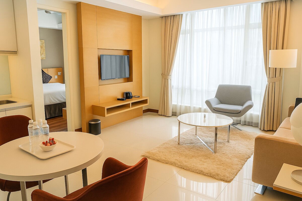 A hotel room with a bed, a TV mounted on the wall, a seating area with a table and chairs, and a large window with curtains.