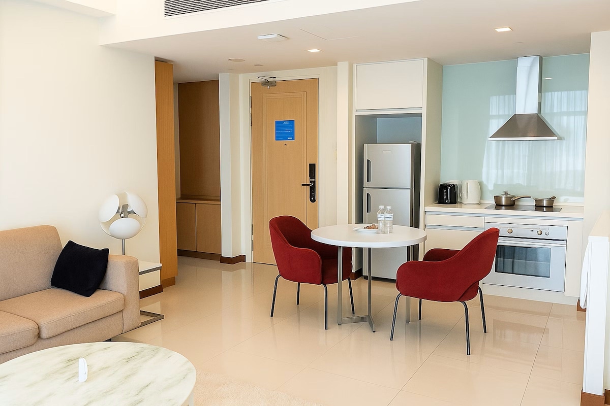 Habitación de hotel moderna con sofá, mesa redonda y dos sillas rojas, además de una cocina equipada con refrigerador, estufa y armarios.