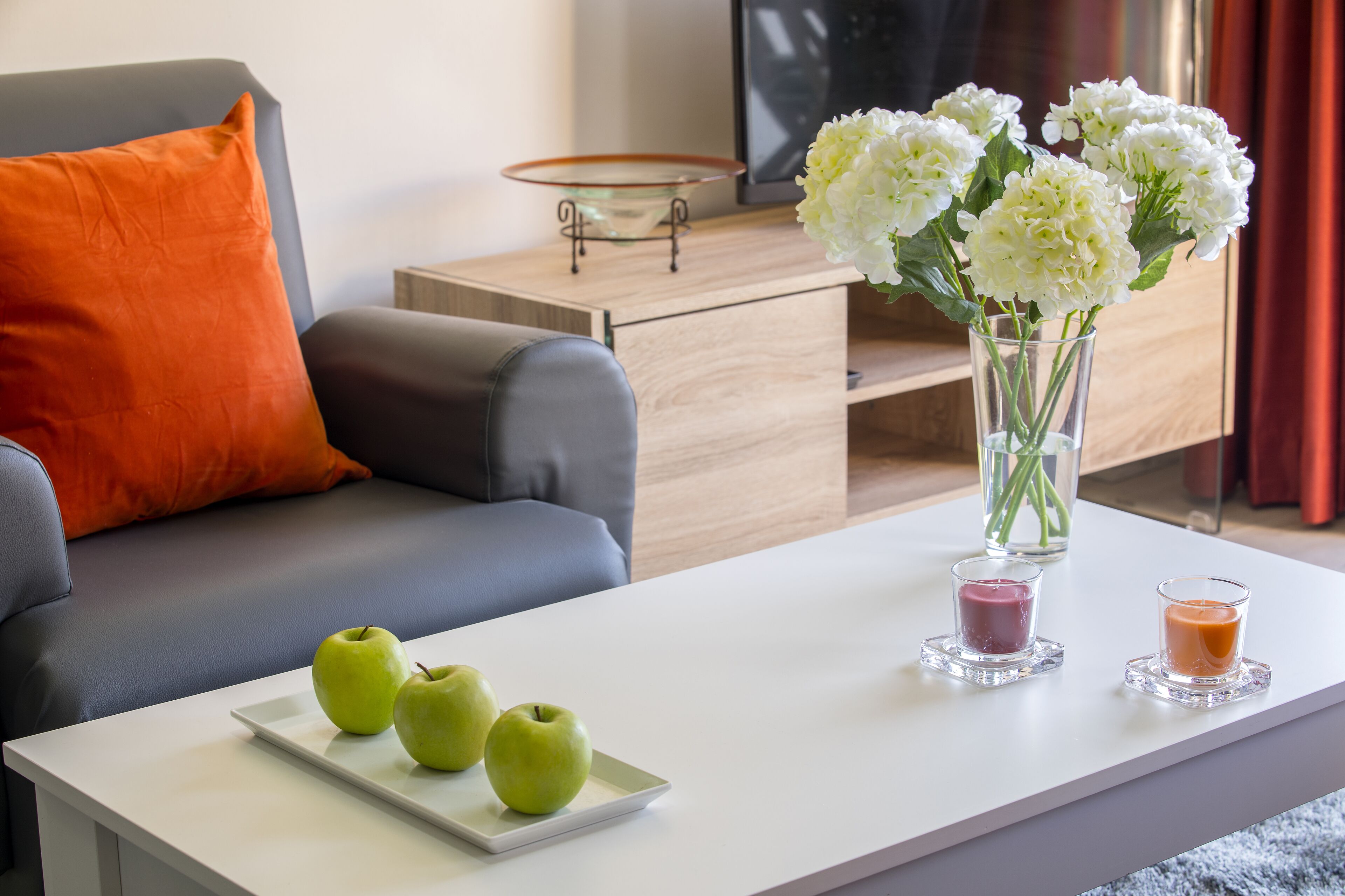 A modern living room with a grey sofa, a white coffee table with green apples, a vase with white flowers, and two candles.
