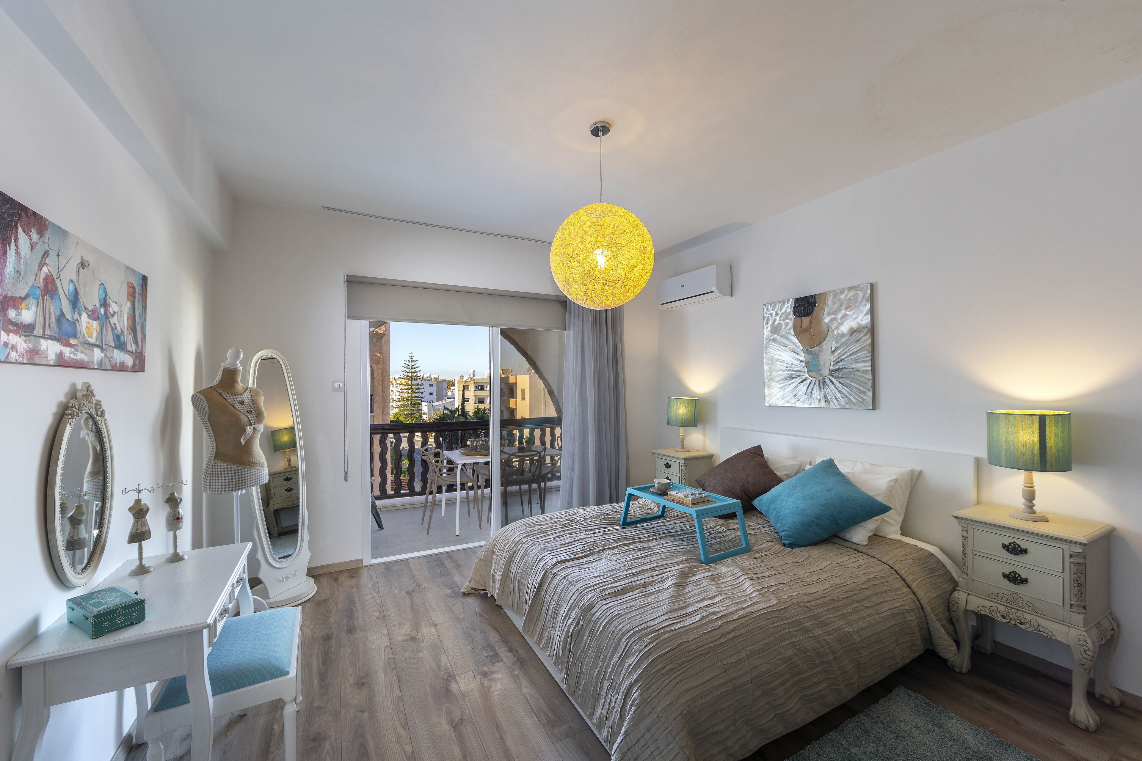 Une chambre avec un grand lit, un bureau, une chaise, une commode et un balcon donnant sur des immeubles.