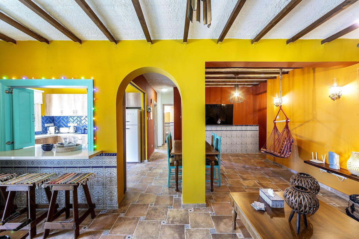 A bright yellow interior with a kitchen area, a dining table with chairs, and a hanging light fixture.