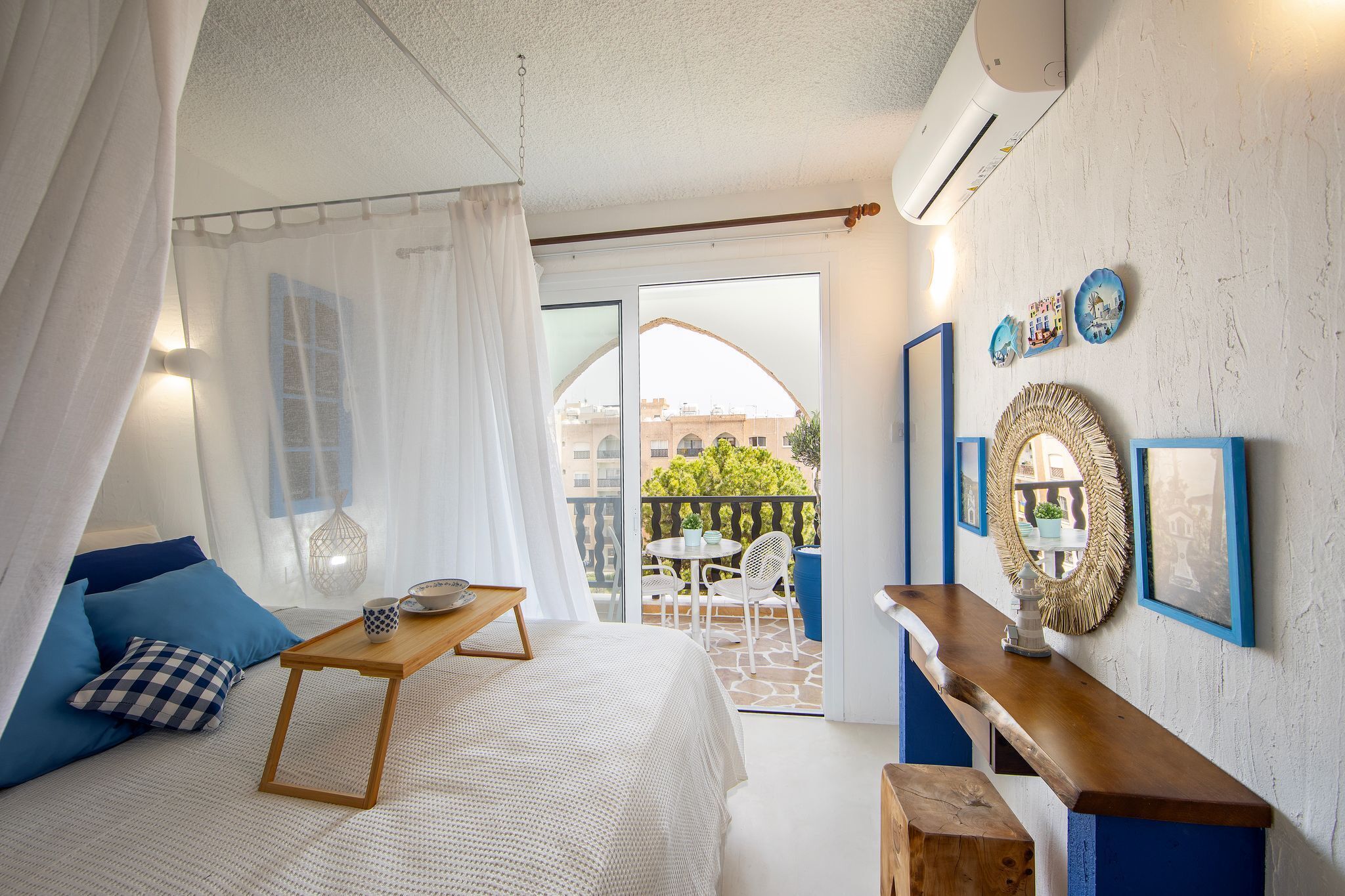 A bedroom with a bed, a wooden side table, a balcony with a table and chairs, and a view of buildings and greenery.