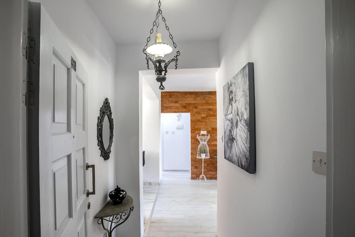 A hallway with white walls, a hanging chandelier, a door with a number plate, a mirror, a coat rack, and a framed picture.