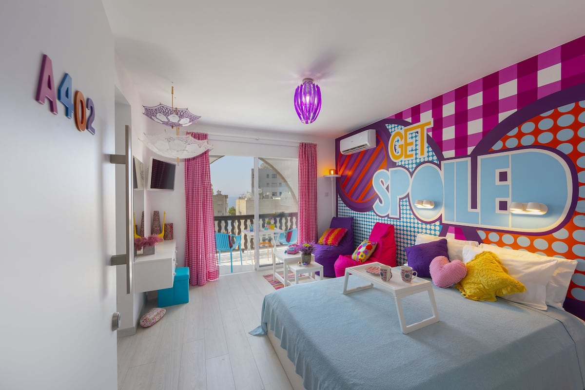 A brightly colored bedroom with a bed, a sofa, and a balcony with a view.