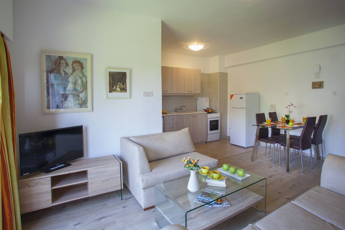 A modern living room with a flat-screen TV, a sofa, a glass coffee table, and a dining area with chairs and a table.