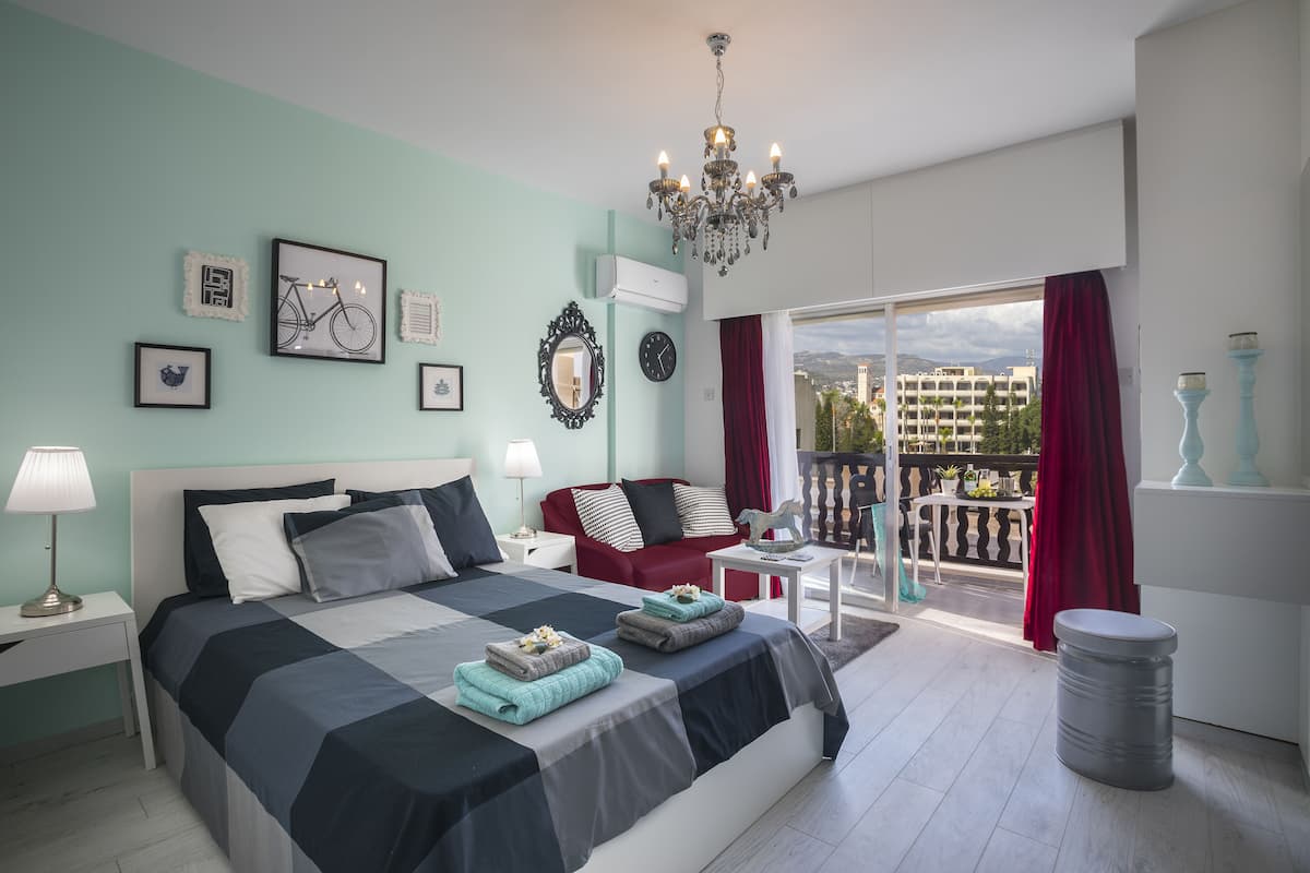 A modern bedroom with a large bed, a red sofa, a balcony with a view of buildings, and a chandelier.