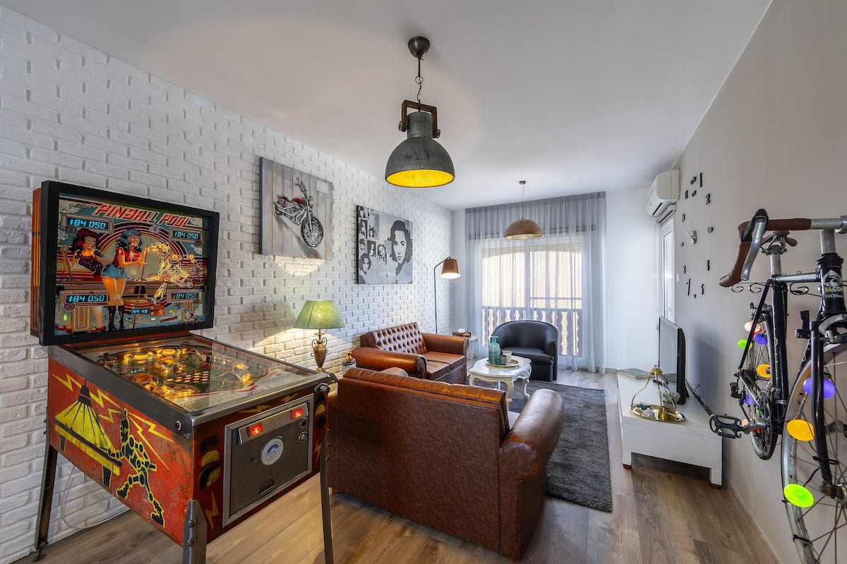 A living room with a pinball machine, leather sofa, and a bicycle.
