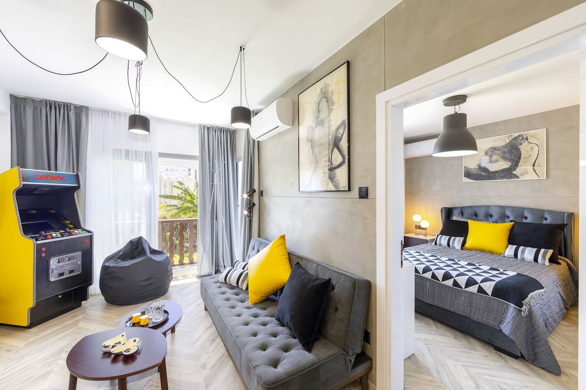 A modern hotel room with a sofa, a bed, a gaming console, and a balcony with curtains.