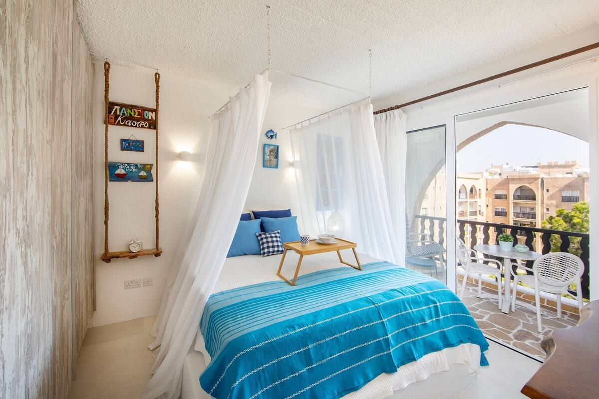 A bedroom with a bed, a bedside table, a hanging sign, and a balcony with chairs and a table.