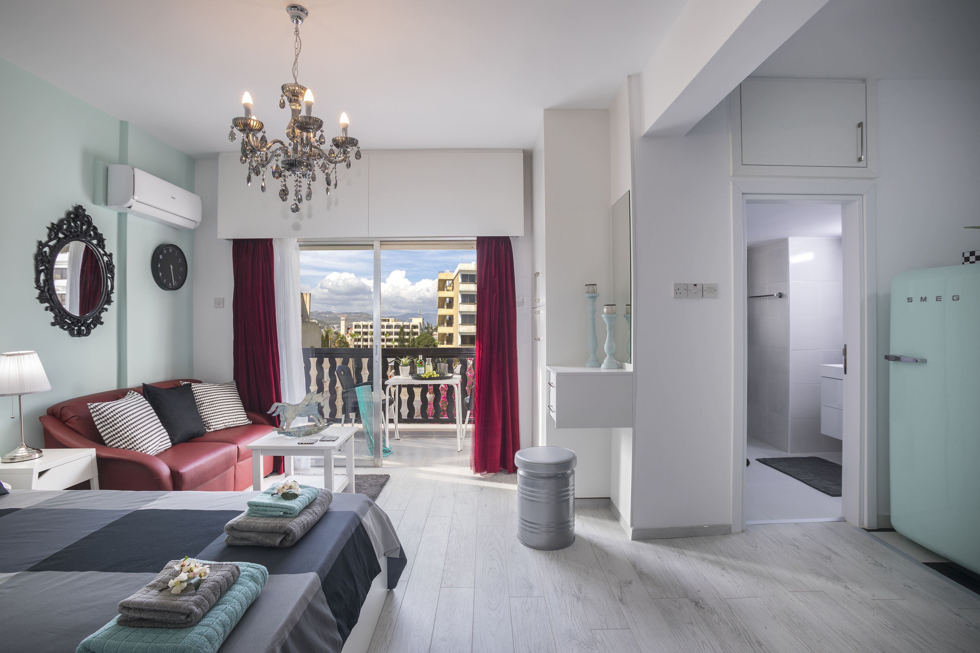 Une chambre d’hôtel moderne avec un lit, un canapé en cuir rouge, un lustre et un balcon agrémenté de rideaux rouges.