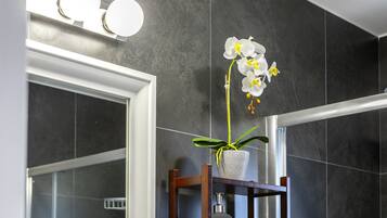 A bathroom with dark tiles, a mirror, a wooden shelf with a potted plant, and a hand soap dispenser.