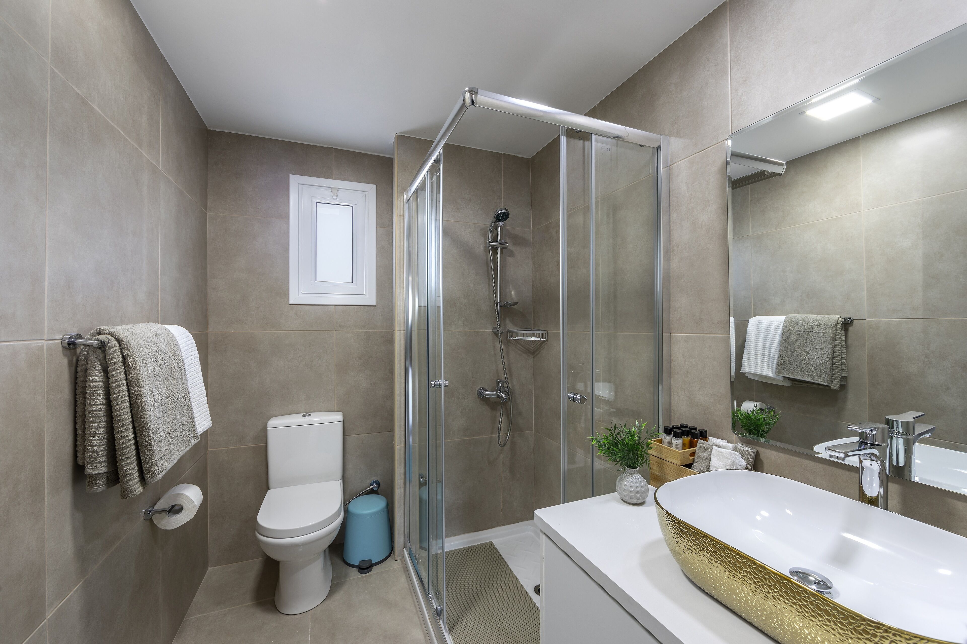 Un baño moderno con ducha envuelta en vidrio, inodoro blanco, lavamanos con lavabo estampado en dorado y un espejo sobre el lavamanos.