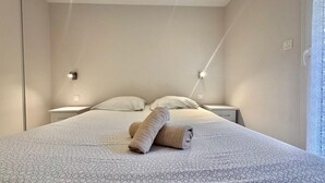 A neatly made bed with a patterned bedspread, two pillows, and a rolled-up towel on the bed. There are two bedside tables with lamps, and a window with a white curtain.
