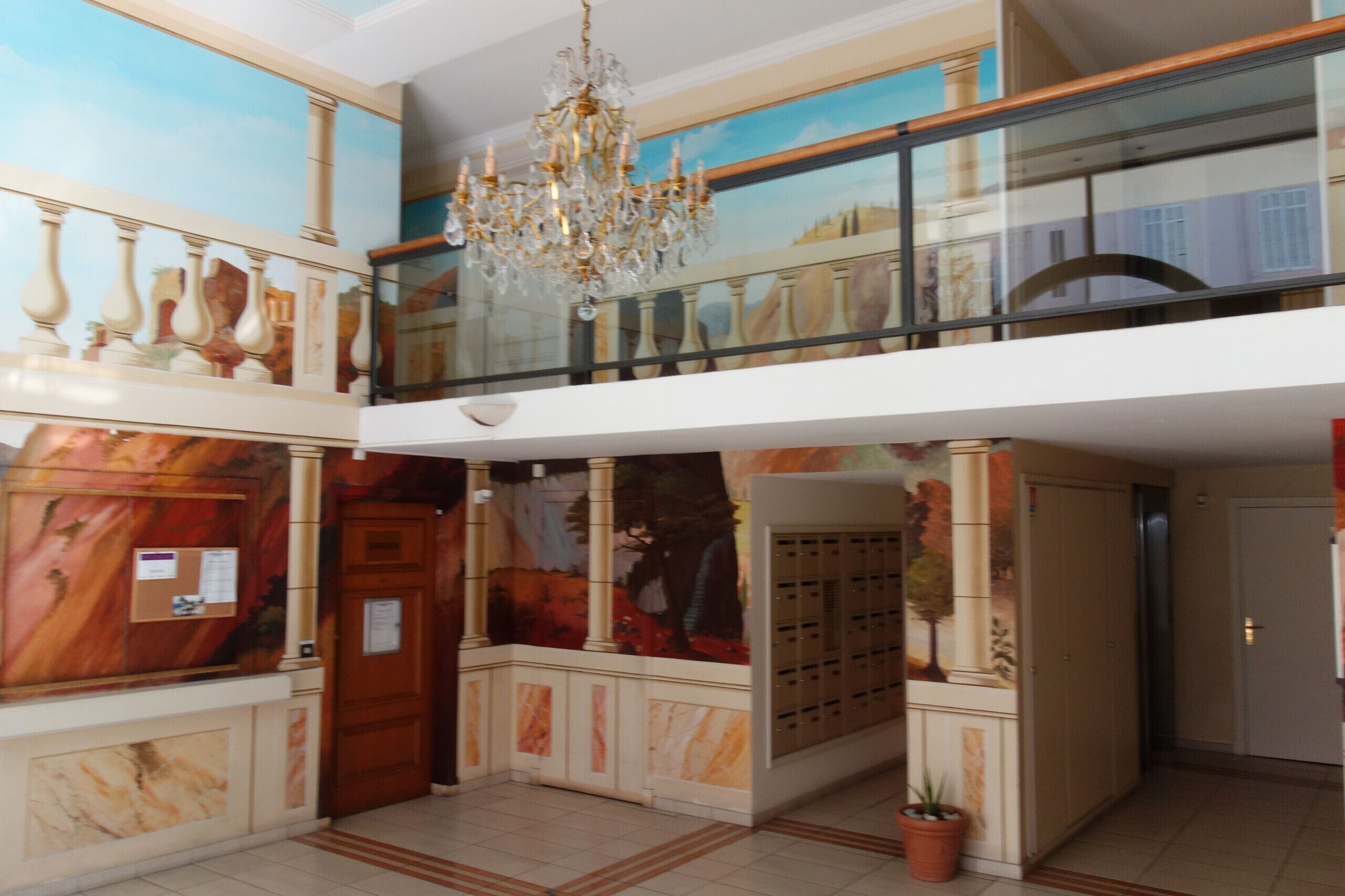 A hotel lobby with a marble reception desk, a chandelier, a glass railing balcony, and a mural on the wall.