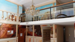 A hotel lobby with a marble reception desk, a chandelier, a glass railing balcony, and a mural on the wall.