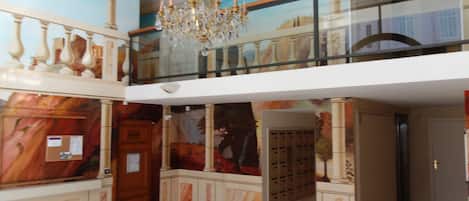 Un hall d’hôtel doté d’un comptoir de réception en marbre, d’un lustre, d’un balcon avec une balustrade en verre et d’une fresque murale.