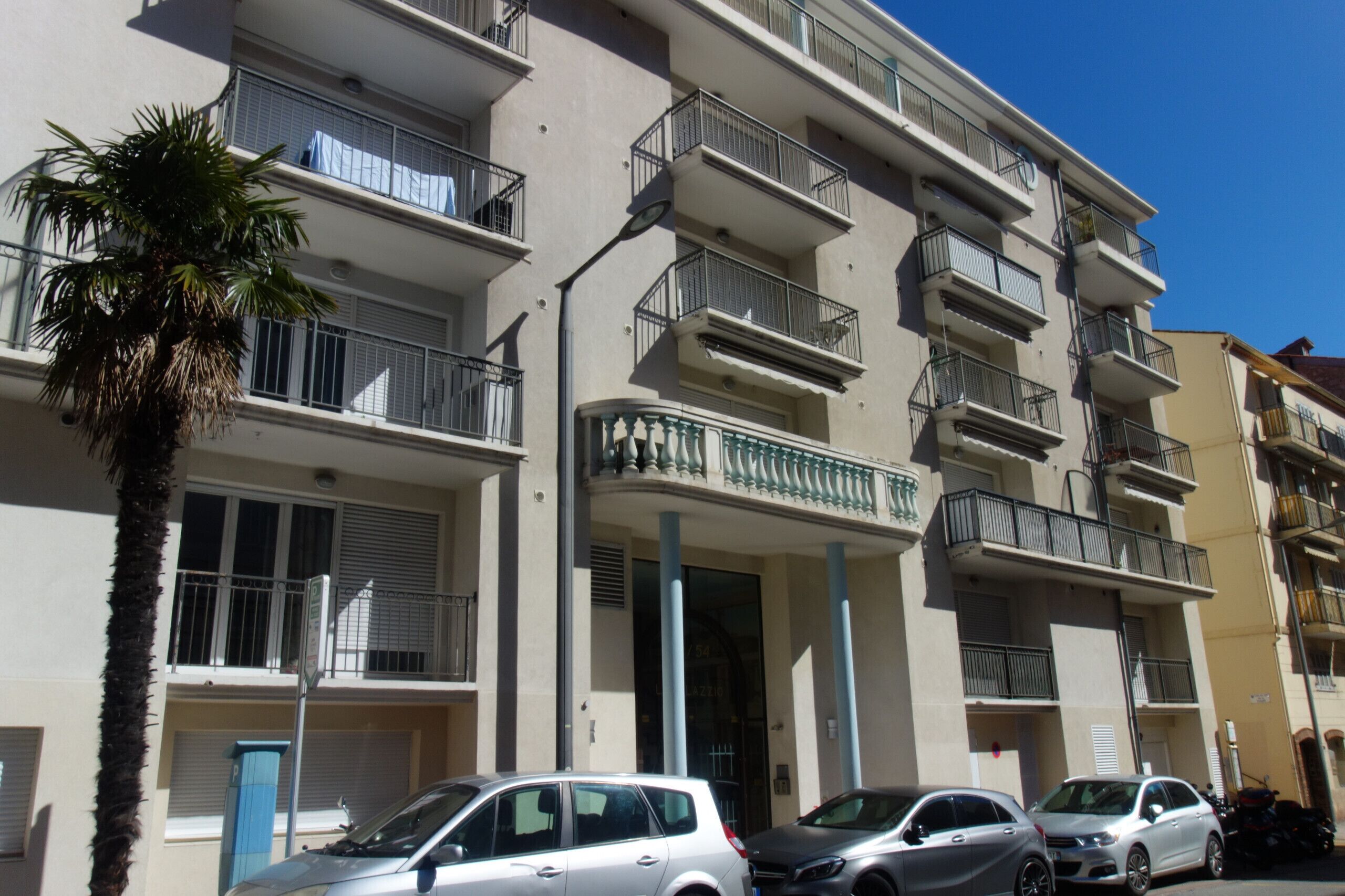 Un immeuble résidentiel à plusieurs étages, doté de balcons, avec des voitures garées et un palmier.