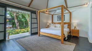 A bedroom with a wooden four-poster bed, a nightstand with a lamp, and a view of a tropical garden through a glass door. - The Aryana Estate (Seminyak)
