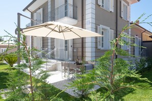 A multi-story building with a balcony, outdoor seating area with a table and chairs, and a large umbrella providing shade. - Villa Maestrale (Camaiore)