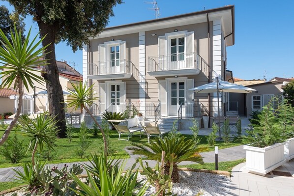 A two-story building with a balcony, surrounded by a garden with various plants and a patio area with outdoor furniture. - Villa Maestrale (Camaiore)