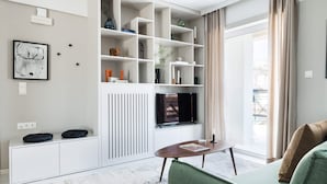 Een moderne woonkamer met een witte inbouwtelevisie meubel, een groene bank en een groot raam met gordijnen.