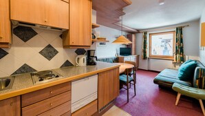 A compact kitchen with wooden cabinets, a built-in dishwasher, and a dining area with a small table and chairs. - Appartement Christine by Skinetworks (Sankt Anton am Arlberg)