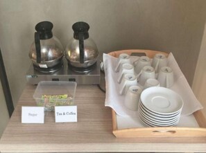 A coffee station with two coffee makers, a sugar container, tea & coffee mugs, and stacked white plates. - Casa Living Setiabudi Jakarta (Jakarta)